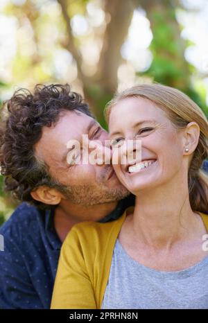 Dandole un grande bacio sulla guancia, un marito affettuoso e una moglie in piedi all'aperto Foto Stock