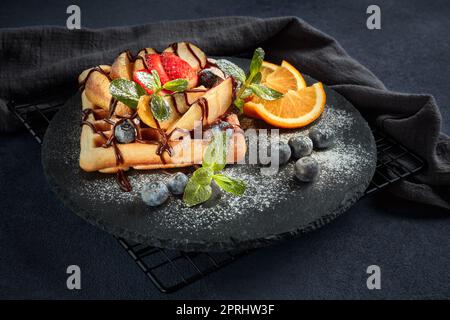 Deliziosi waffle con pera caramellata, frutti di bosco, menta, zucchero a velo e salsa dolce sul piano di servizio nero su sfondo marrone scuro. Dessert. Servizio Foto Stock