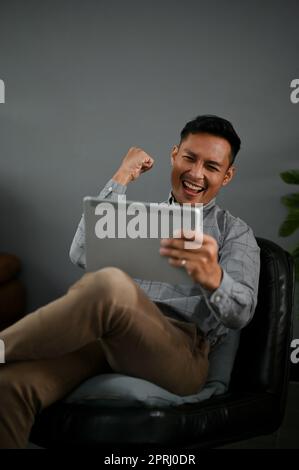 Ritratto di un allegro uomo asiatico millenario in abiti casual guardando il suo schermo tablet e celebrando il suo successo. Foto Stock