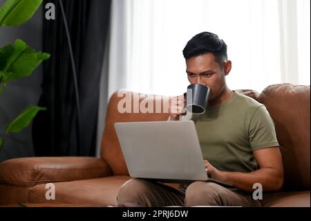 L'astuto uomo asiatico millenario sorseggiando un caffè e utilizzando il suo computer portatile su un divano nel soggiorno, si concentra sulle sue attività sul notebook. lavoro da casa c Foto Stock