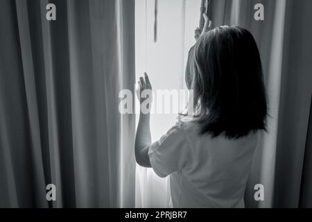 Vista posteriore di una donna che tiene una tenda e guarda fuori dalla camera da letto. Donna depressa e stressata. Concetto di giornata mondiale di salute mentale. Premiati per la salute mentale. Donna premurosa in una stanza buia. Donna infelice. Foto Stock