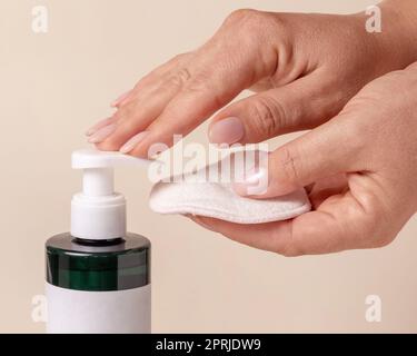 Una mano donna pompando liquido su un tampone di rimozione trucco da una bottiglia cosmetica su primo piano beige. Confezione di prodotti di bellezza per la cura della pelle, composizione minima Foto Stock