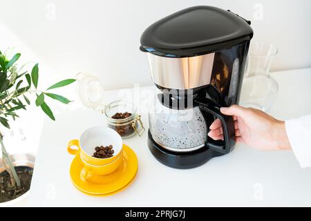 Bella mattina, la ragazza sta aspettando il suo caffè. Una macchina per caffè antigoccia automatica è dotata di una tazza gialla su un tavolo bianco. Cucina elettrica smal Foto Stock