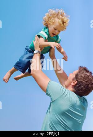 Il padre pieno di divertimento. Un padre che dondola il figlio del bambino nell'aria contro uno sfondo del cielo Foto Stock