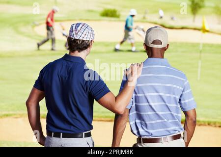 In attesa di salire sul buco. Ripresa da dietro di due amici che guardano un gruppo di golfisti in lontananza Foto Stock