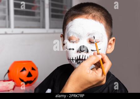 Ritratto di donna che applica spazzola di colore al viso del bambino per il partito a casa, famiglia madre asiatica e figlio del capretto che ottiene trucco halloween pittura del viso Look li Foto Stock