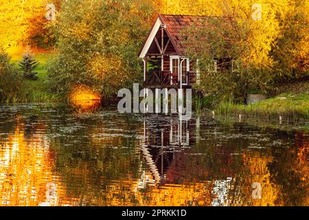 Cabina vacanza rossa con riflesso nello stagno durante il tramonto. Cottage in legno, sauna sulla riva. Piccola casa vicino all'acqua. Edificio circondato dall'autunno Foto Stock