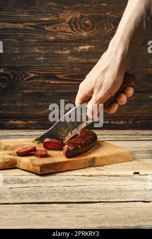 A mano con coltello taglio di manzo affumicato e salsiccia di cervo su fette di tagliere di legno Foto Stock