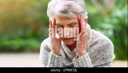 Queste emicranie mi irritano. una donna anziana stressata seduta su una panchina e tenendo la testa in disagio all'esterno in un parco Foto Stock