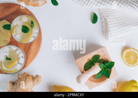 Due bicchieri con limonata e ingredienti per cucinare. Zenzero, limone, menta, ghiaccio su superficie bianca. Vassoio di legno, mortaio e pestello. Disposizione piatta. Spazio di copia Foto Stock