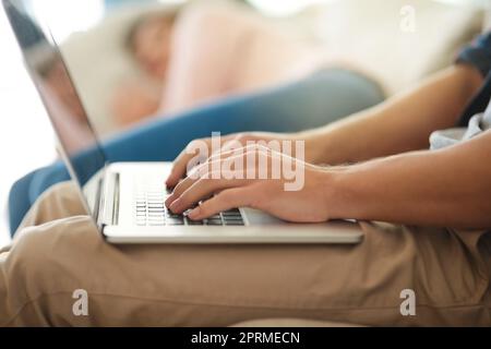 Cercando di raggiungere questo equilibrio nella vita lavorativa. Un uomo non identificabile che lavora sul suo notebook a casa mentre sua moglie si trova sul divano accanto a lui. Foto Stock