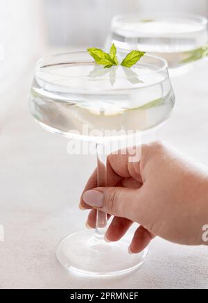 Tenere a mano un cocktail trasparente in un bicchiere coupé decorato con foglie di menta da vicino Foto Stock