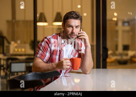 Ritratto di un uomo bearded attraente che indossa abiti casual seduto su un divano in soggiorno, utilizzando il telefono cellulare. Bel testo uomo millenario m Foto Stock