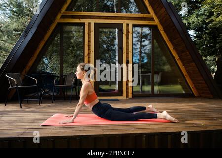 Giovane donna fa serpente o cobra posa all'aperto su tappeto fitness sulla terrazza del suo bungalow Foto Stock