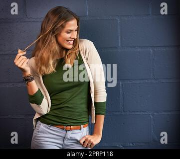 Solo un po 'di timidezza può essere accattivante. Una giovane donna che gioca con i suoi capelli contro uno sfondo muro di mattoni. Foto Stock