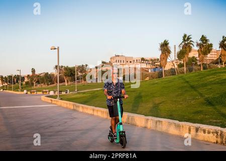 Giovane a cuore uomo di 50s anni che usa scooter elettrico lungo passeggiata / Beach percorso verso il tramonto su scooter Foto Stock