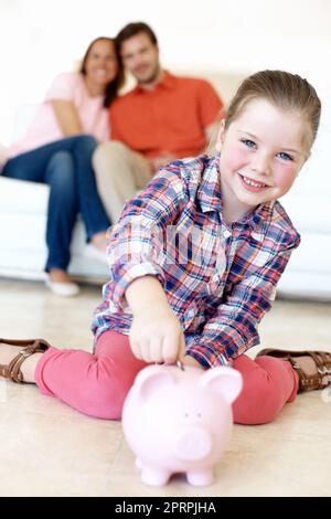 Insegnare ai nostri figli a risparmiare. Una ragazza giovane mette i soldi in un piggybank mentre i suoi genitori guardano. Foto Stock