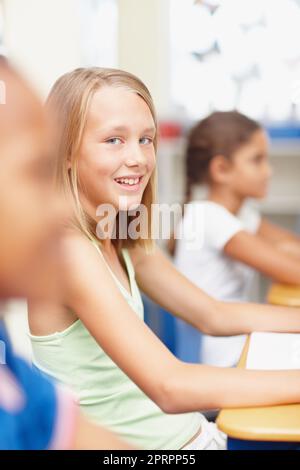Sentirsi fiduciosi nella mia classe. Giovane studentessa bionda che guarda la macchina fotografica mentre si siede alla sua scrivania in classe - copyspace. Foto Stock
