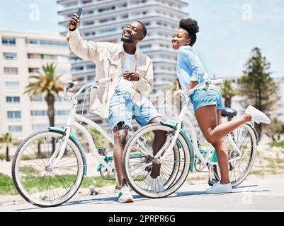 Selfie, bicicletta e appuntamento con una coppia in bicicletta su una passeggiata durante l'estate con amore, romanticismo e affetto. Bicicletta, fotografia e divertimento con una donna e un uomo neri fuori e una città sullo sfondo Foto Stock