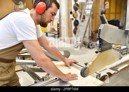 giovane falegname in abiti da lavoro lavora in falegnameria su una levigatrice - indumenti da lavoro con protezione per le orecchie Foto Stock