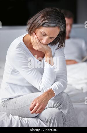 Sentendosi blu. Una donna matura che sembra triste mentre si siede sul suo letto con il marito sullo sfondo. Foto Stock