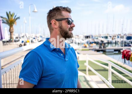 Contenuto bel giovane uomo bearded in occhiali da sole visitando yacht club: Hippster ragazzo all'aperto Foto Stock