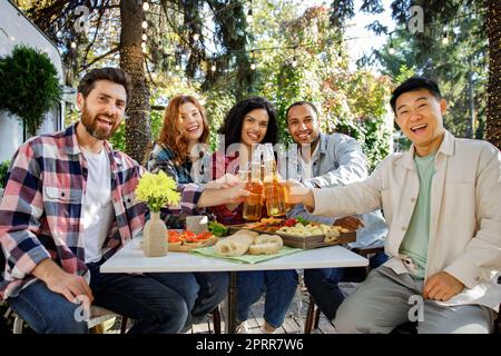Un'allegra azienda multinazionale si diverte a fare un picnic. Amici Multisex seduti al tavolo all'aperto per celebrare il mangiare e bere bevande Foto Stock
