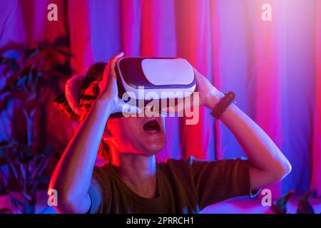 Donna asiatica indossa il casco VR eccitato emotivo a casa sfondo viola e blu, femmina che sperimenta la realtà virtuale occhiali di protezione vivere la realtà, Foto Stock