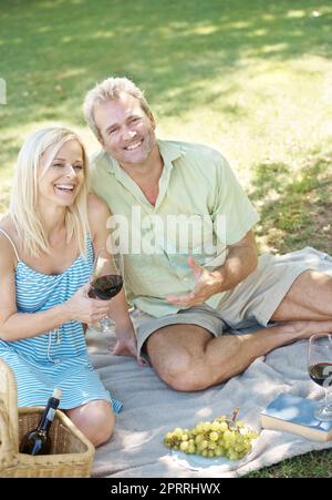 Buon vino e grande compagnia. Un marito e una moglie felici che si godono un bicchiere di vino mentre si ha un pic-nic all'aperto in un parco in una giornata estiva. Foto Stock