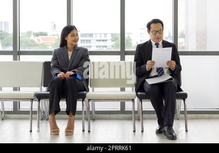 Giovane donna guarda e sorride un uomo seduto più lontano. Uomini d'affari asiatici in attesa di un colloquio di lavoro seduti separati. Concetto di nuovo ufficio normale durante la pandemia di coronavirus Foto Stock