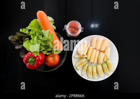 Una ciotola di vetro con una varietà di frutta e verdura è posizionata sul bancone della cucina. Peperoni, pomodori, carote e lattuga sono in un recipiente di vetro. Un piatto con mela a fette è stato posto accanto ad esso. Foto Stock