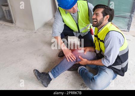 Il giovane uomo caucasico in giubbotto di sicurezza gli afferrò il ginocchio e urla nel dolore. A causa di lesioni in cantiere. Il suo amico asiatico viene e lo aiuta a stare in piedi. Foto Stock