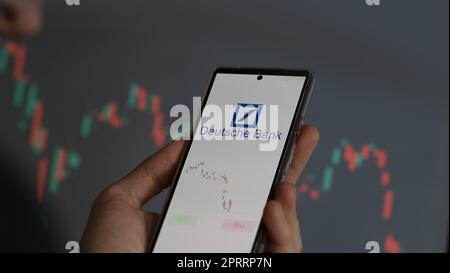 Un investitore che analizza le azioni di Deutsche Bank su uno schermo del telefono, alcune tabelle di scambio di azioni sono sullo sfondo. Foto Stock