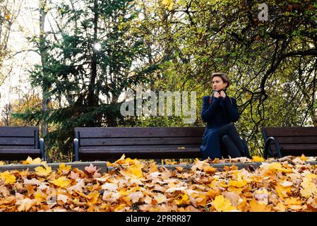 Una donna si avvolge in un cappotto seduto su una panchina in un parco autunnale Foto Stock