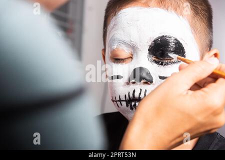 Madre asiatica e figlio di capretto ottenere trucco haloween pittura faccia assomiglia a fantasma, ritratto della donna che applica il pennello di colore al viso del bambino per partito, felice Foto Stock