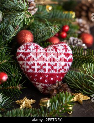 Cuore a maglia rossa tra decorazioni natalizie, rami di abete e coni di pino da vicino Foto Stock