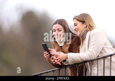 Due amici felici che controllano il telefono in inverno in un balcone Foto Stock