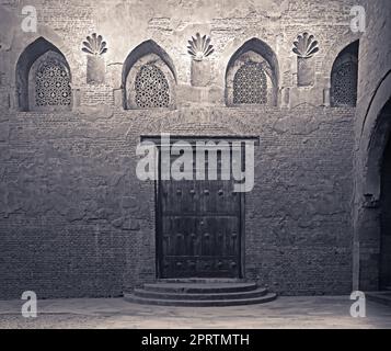 Invecchiato in legno porta a spiovente, arcuata forata finestra di stucco decorato con motivi floreali e tre passaggi su pietra muro in mattoni, Ibn Tulun Mosque, Ca Foto Stock