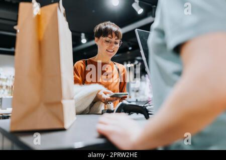 Donna matura sorridente che effettua il pagamento tramite smartphone al banco cassa nel negozio di elettronica Foto Stock