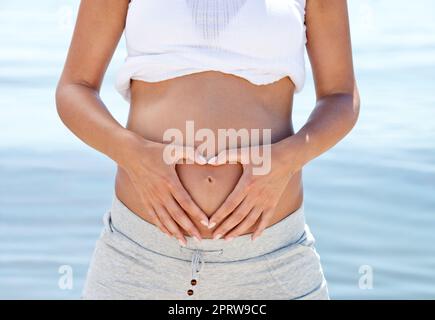 Ora di tum. Una donna incinta che fa una forma del cuore intorno al suo ventre su uno sfondo dell'oceano. Foto Stock