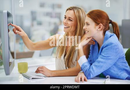 Guardate quanto può essere facile. Una collega attraente che mostra alla sua collega qualcosa di divertente sul suo computer. Foto Stock