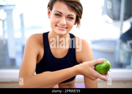 Mangiare sano la mantiene bella. Una giovane donna sportiva seduta sul pavimento di una palestra che mangia una mela. Foto Stock