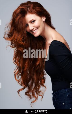 La vita non è perfetta, ma almeno i miei capelli lo sono. Studio shot di una giovane donna con bei capelli rossi che si posano su uno sfondo grigio. Foto Stock