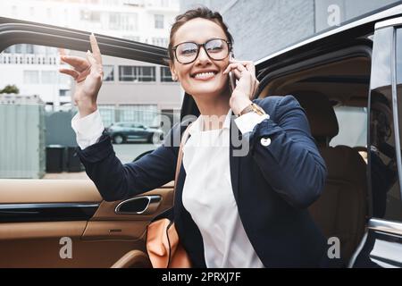 Im proprio qui. una giovane donna d'affari fiduciosa che esce da un'auto mentre tiene un cellulare e solleva la mano fuori durante il giorno. Foto Stock