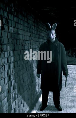 uomo misterioso con un lungo cappotto e maschera di coniglio vicino ad un muro, un coltello in mano Foto Stock