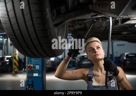 Un giovane artigiano esegue lavori di riparazione sotto l'auto sollevata sull'ascensore Foto Stock