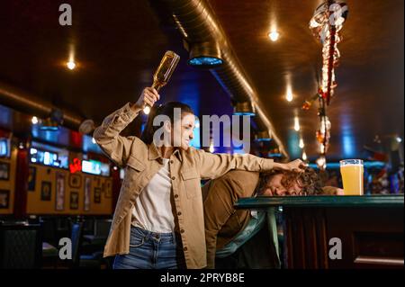 Tifosi infelici e arrabbiati che combattono allo sport bar. Pazzo pazzo donna cercando di rompere la bottiglia di birra sulla testa maschile. Tifosi di calcio problematici. Le persone sono emotive e cattive Foto Stock