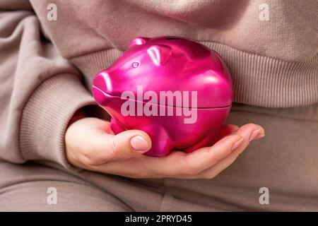Mano donna che tiene in primo piano rosa brillante scatola di denaro maiale. Economia, budget e risparmio. Foto Stock