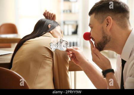 Uomo con il naso di clown che attacca il pesce di carta alla schiena della collega mentre dorme in ufficio. Scherzo divertente Foto Stock