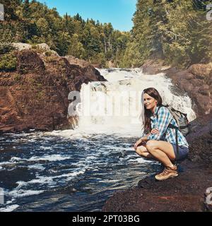 La natura, fonte di vita. una giovane donna attraente accovacciata accanto ad un fiume roccioso e cascata. Foto Stock